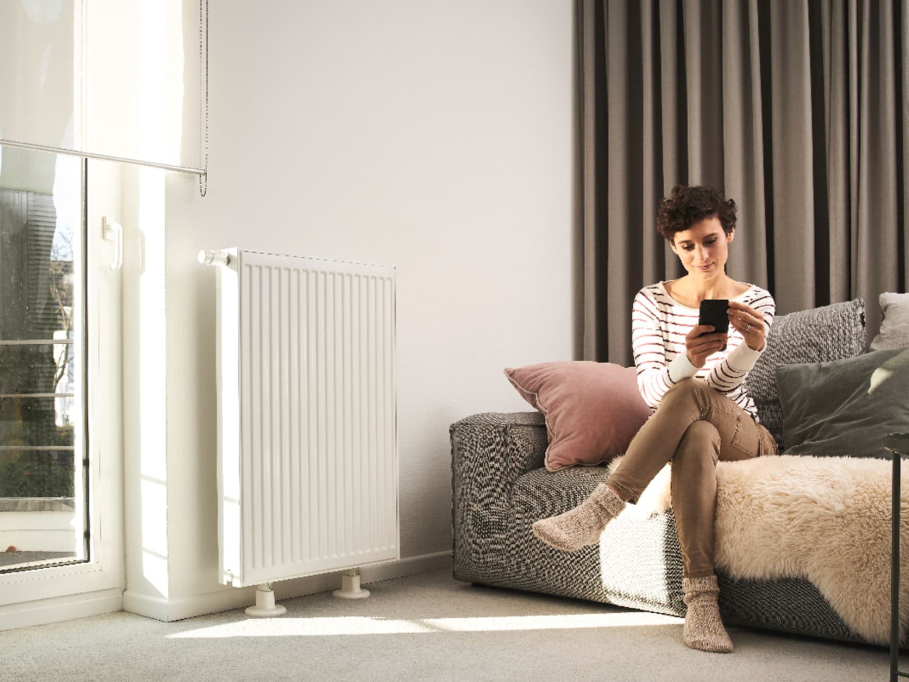 Woman sits on sofa using a smartphone. Sofa is in front of a window and beside a radiator