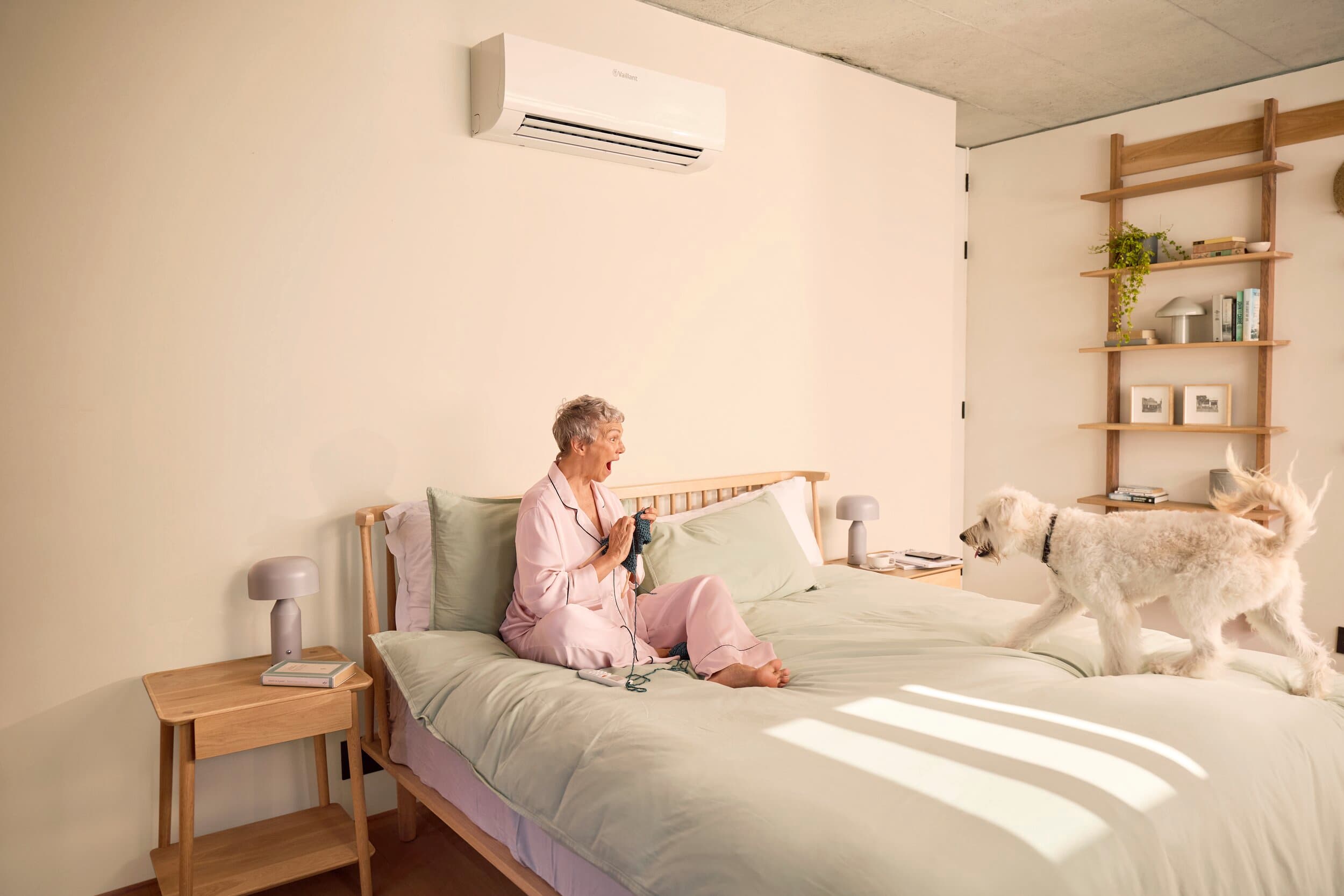 A woman is sitting in her bed as her dog comes over to her