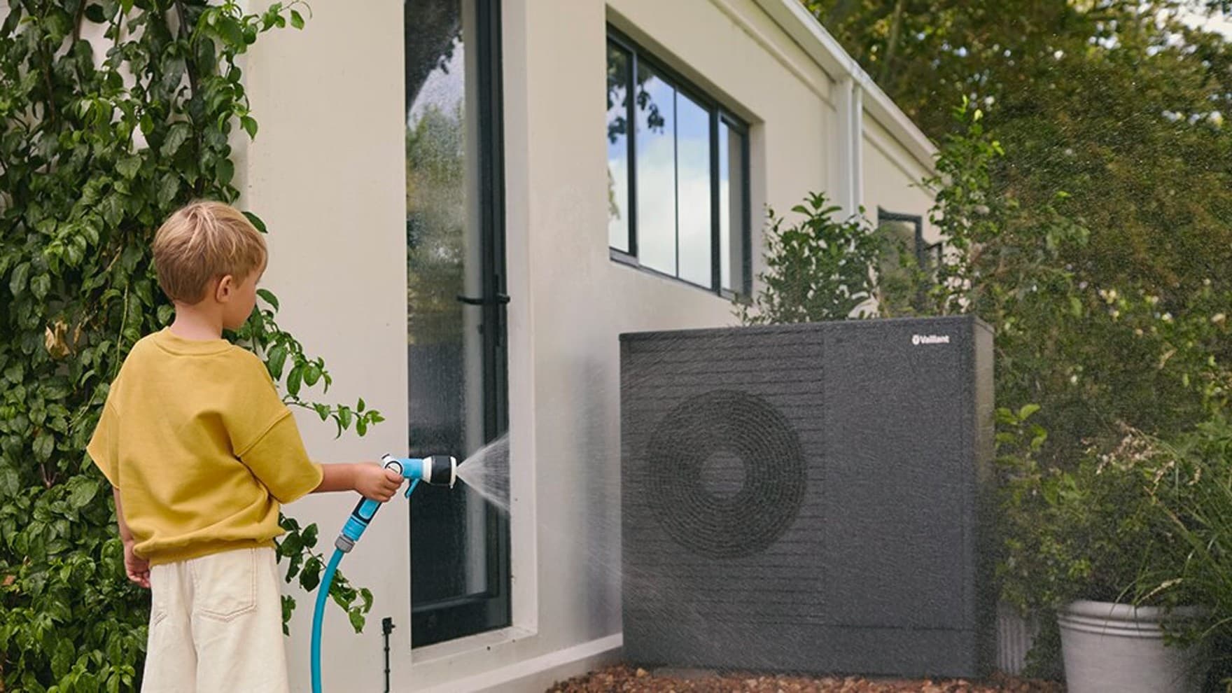 A boy sprays water onto the heat pump with the garden hose