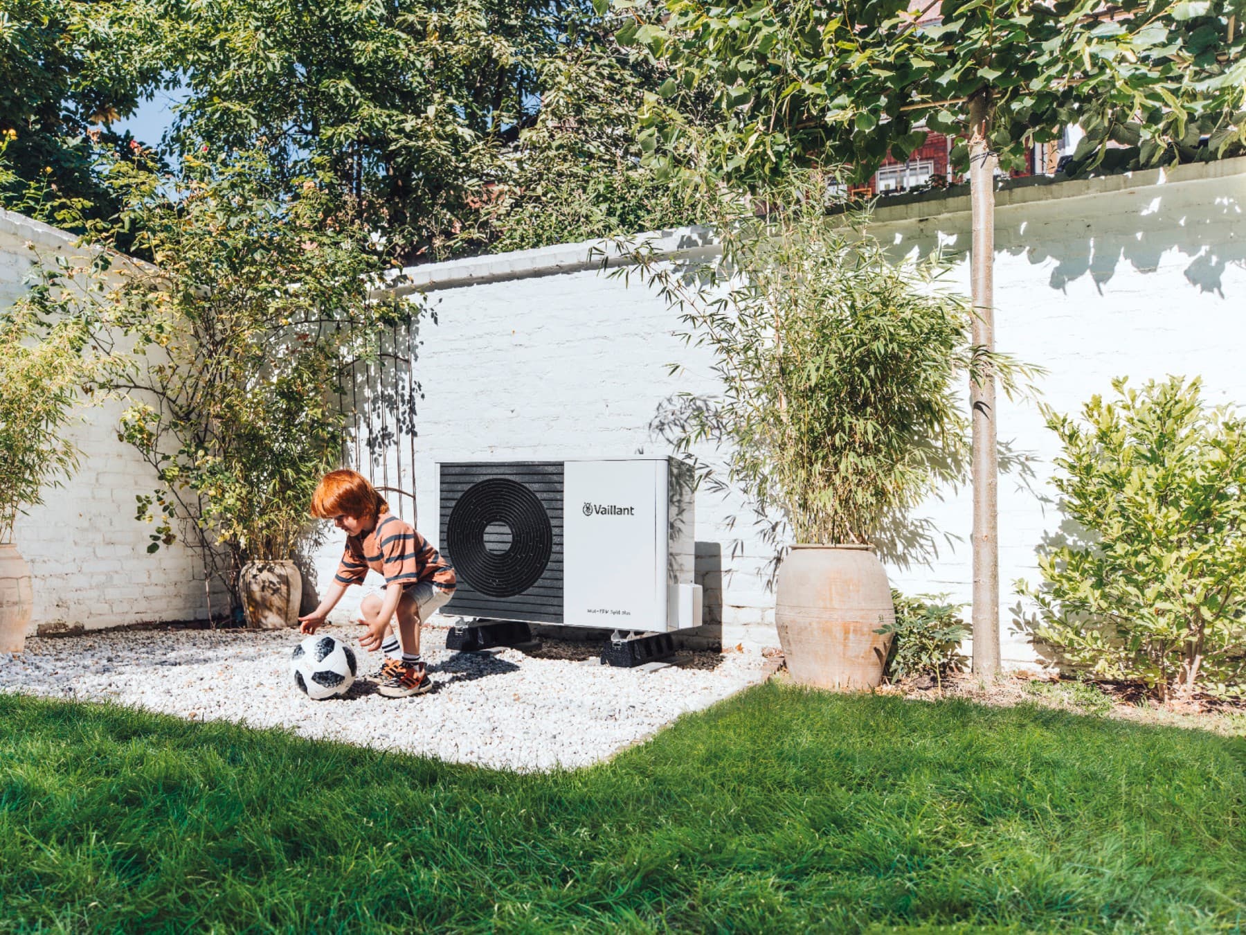 Enfant jouant au football dans le jardin devant une pompe à chaleur