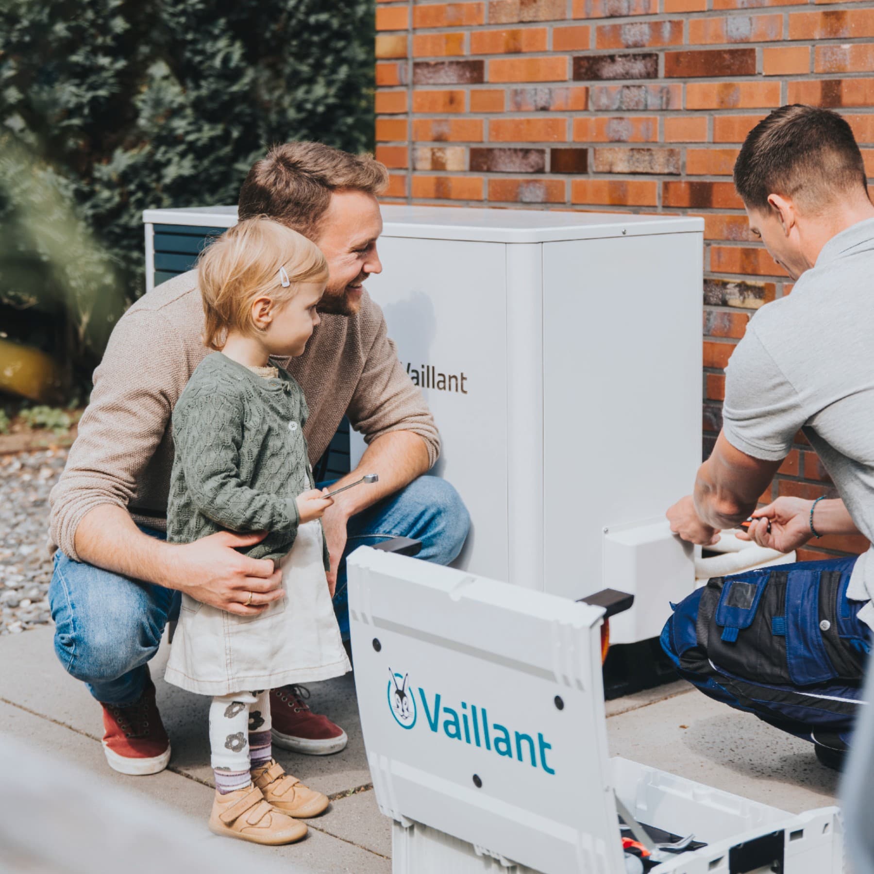 A man with his daughter and a plumber standing in front of a heat pump outdoor unit.