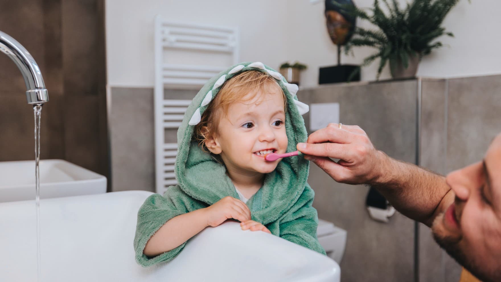 Father brushes child's teeth with a heated towel rail in the background