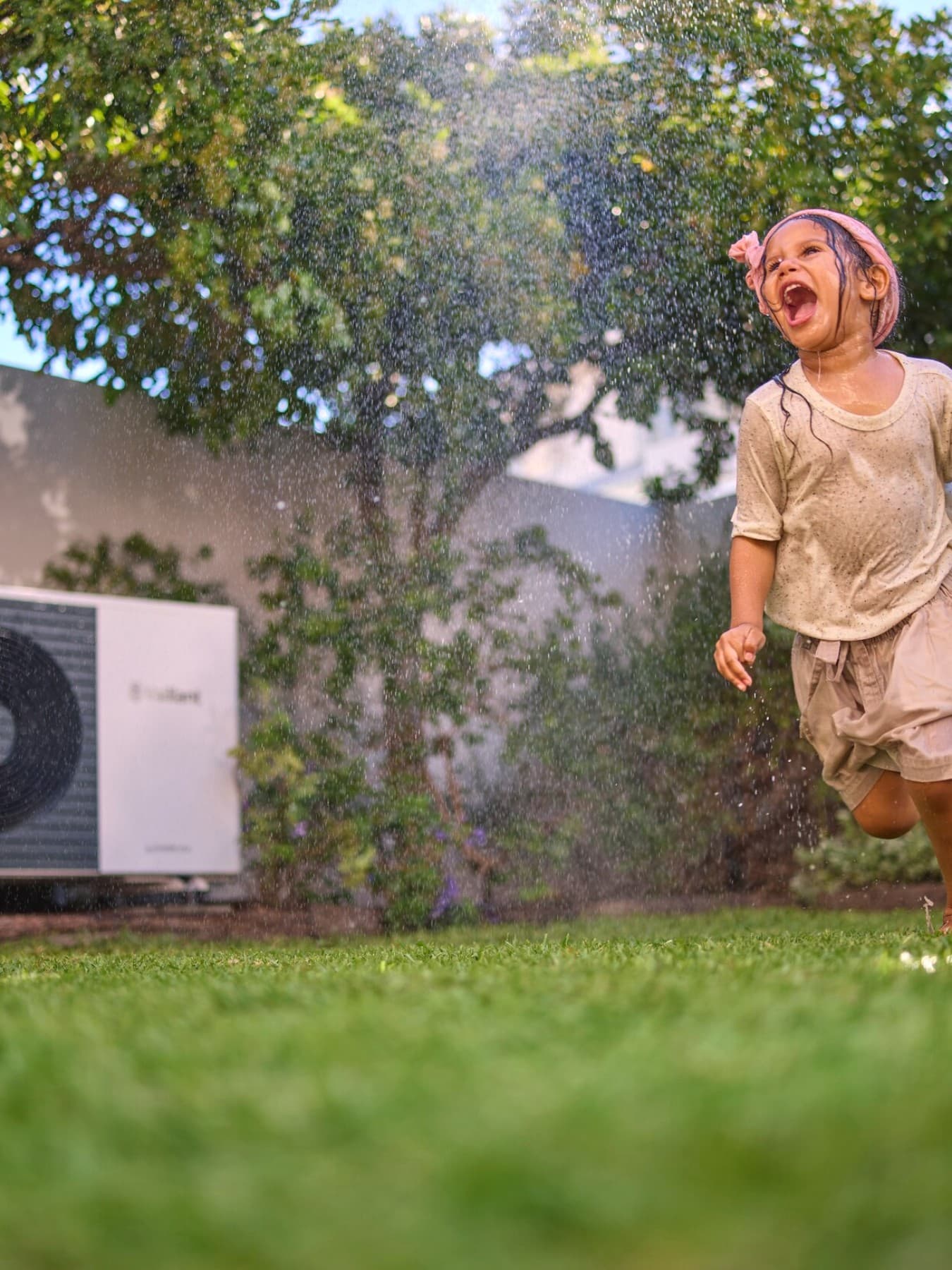 A girl is running through the sprinkler. A heat pump can be seen in the background.