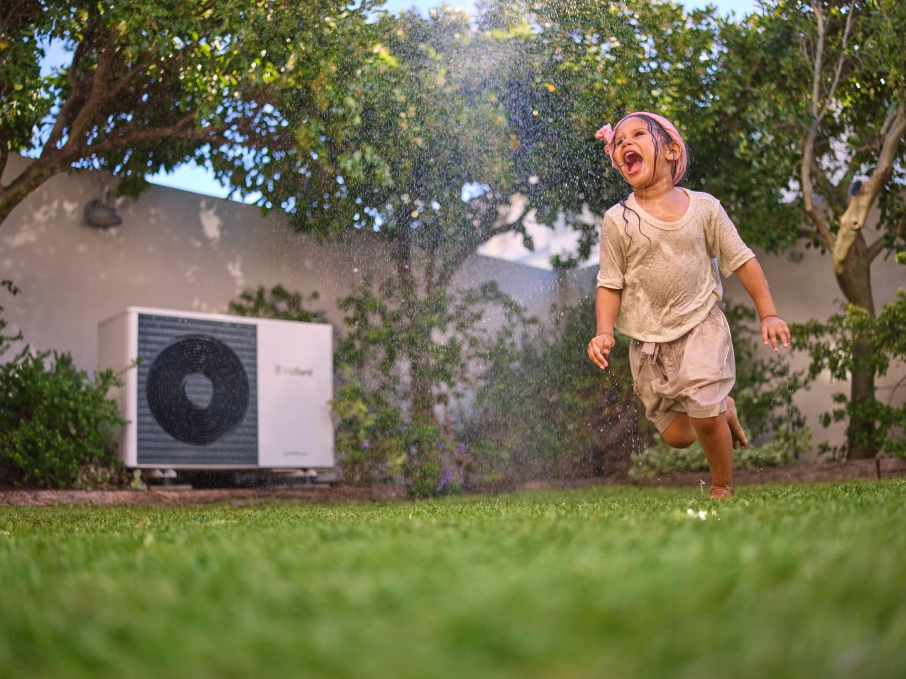 A girl is running through the sprinkler. A heat pump can be seen in the background.