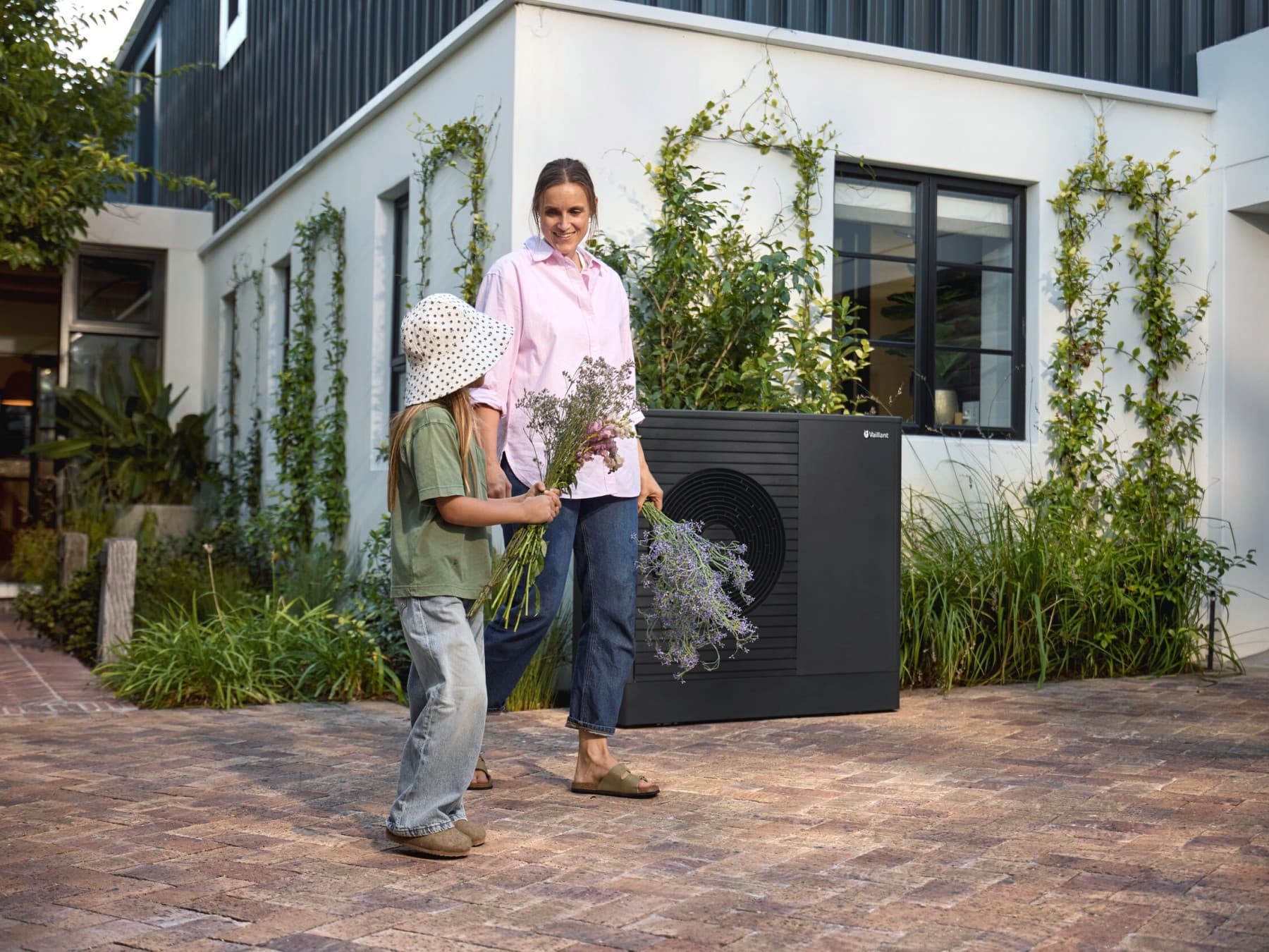 A woman and child stand in front of a house with a heat pump, holding flowers in their hands.