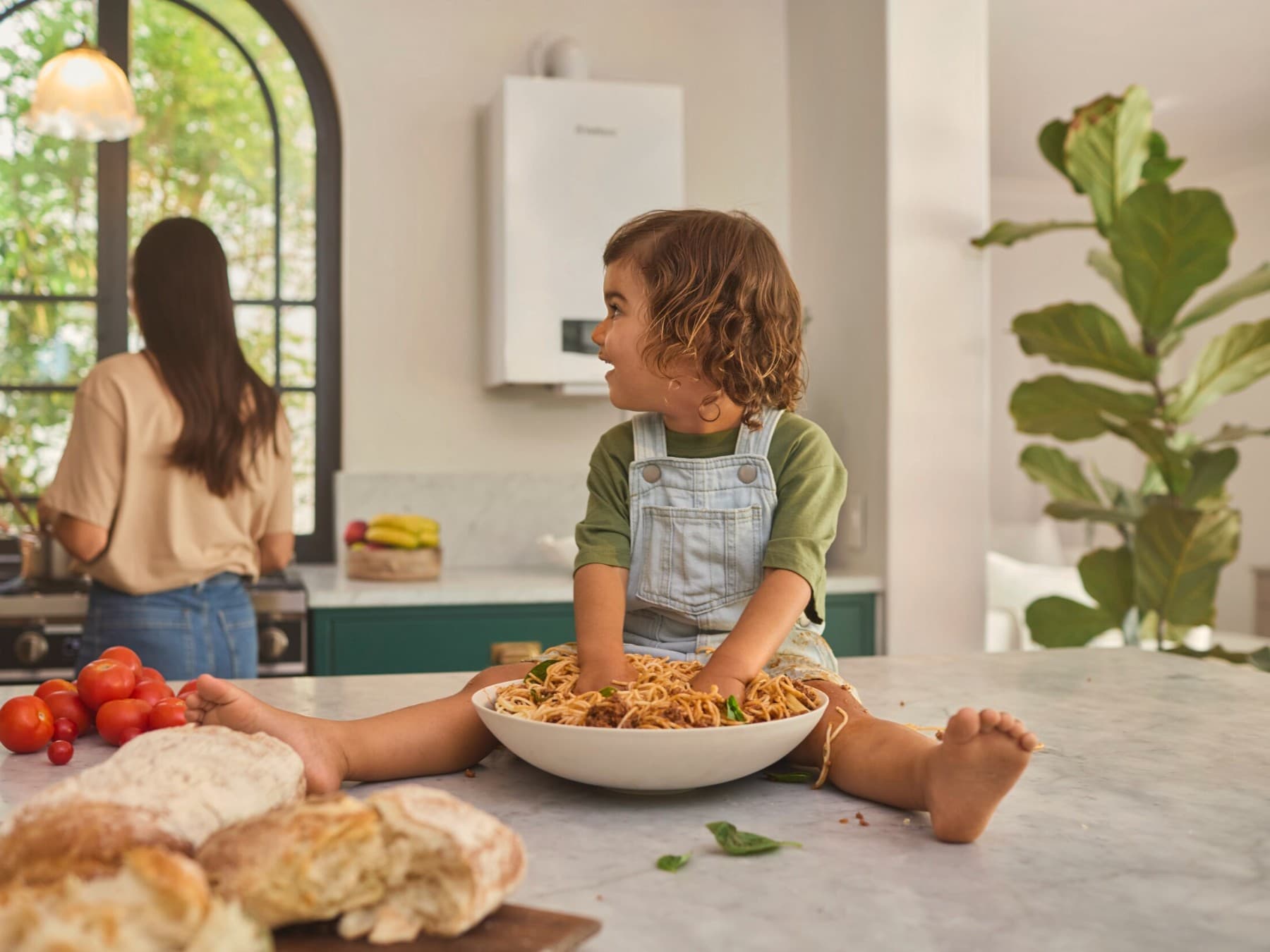 Un enfant est assis sur le comptoir pendant que ses parents cuisinent. Un chauffe-eau Vaillant fournit de l'eau chaude fraîche.